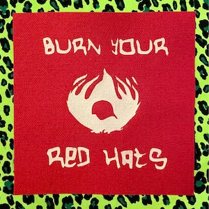 May include: A square red fabric patch featuring the white text BURN YOUR RED HATS printed in a stylized font. Centered between the two lines of text is a white graphic illustration of a flame shape with a small circular cutout in the center.