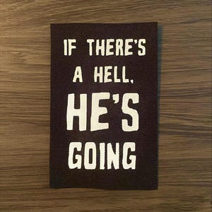 May include: A rectangular black fabric patch with white text that reads "IF THERE'S A HELL, HE'S GOING". The patch is set against a wooden surface.