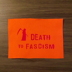 May include: Orange rectangular patch with the words "DEATH TO FASCISM" in red block letters. A red silhouette of the Grim Reaper with a scythe is above the word "DEATH". The patch is made of a textured fabric.