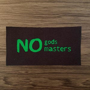 May include: A rectangular patch with the words "NO gods masters" in bright green lettering on a dark brown background. The patch is set against a wooden surface.