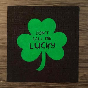 May include: A square, black fabric patch featuring a bright green shamrock. The shamrock has the text "DON'T CALL ME LUCKY" in black, block letters. The patch is likely for clothing or accessories.