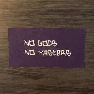 May include: A rectangular purple patch with the white text "NO GODS NO MASTERS". The patch is made of a textured fabric and is displayed on a wooden surface.