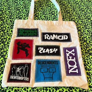 May include: A cream-colored canvas tote bag featuring several rectangular fabric patches sewn onto the front. The patches display band names and logos including Dropkick Murphys, Rancid, The Clash, NOFX, The Interrupters, and the Descendents.