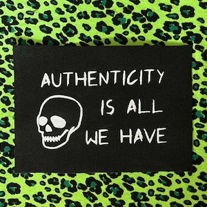 May include: A black rectangular patch with the words "AUTHENTICITY IS ALL WE HAVE" in white, along with a white skull graphic. The patch is set against a vibrant green background with a black leopard print.