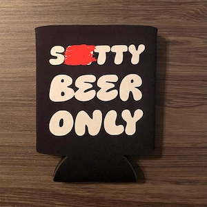 May include: Black beer can cooler with the words "S**tty Beer Only" in white, bold, bubble letters. The word "S**tty" has a red paint stroke over it. The cooler is made of a black, insulating material.