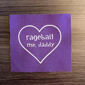 May include: A square, purple fabric patch with a white heart outline. Inside the heart, the words "ragebait me, daddy" are printed in a sans-serif font. The patch is likely for sewing or ironing onto clothing or accessories.