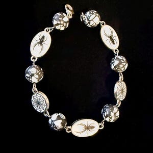 Puede incluir: Una pulsera plateada con cuentas ovaladas y redondas. Las cuentas ovaladas muestran un fondo blanco con un diseño de araña o telaraña negra. Las cuentas redondas tienen un patrón moteado en blanco y negro. La pulsera se abrocha con un cierre.