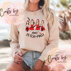 May include: Beige crewneck sweatshirt featuring a graphic of four women in Santa hats and red dresses. The text "MERRY FETCH-MAS!" is below the graphic. The sweatshirt has long sleeves and a relaxed fit.
