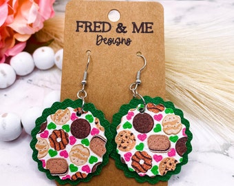May include: Round, layered earrings with a green scalloped edge. The top layer features a white background with various cookie designs, including chocolate, peanut butter, and shortbread. The earrings are attached to a brown card with the text "FRED & ME Designs".