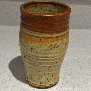 May include: A handmade ceramic vase with a cylindrical shape. The vase features a speckled beige base, transitioning to a band of orange and brown near the top. The rim has a darker brown color. The vase has a rustic, handcrafted appearance.