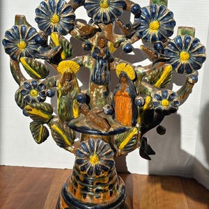 May include: A colorful, handcrafted ceramic tree of life sculpture. The tree features blue and yellow flowers, green leaves, and figures of religious figures. The base is a dark blue and orange color. A unique decorative piece.