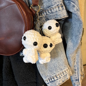 May include: Three white crochet doll keychains with black button eyes hang from a silver keyring. The keychains are attached to a brown leather bag and a denim jacket. The keychains are a handmade craft.