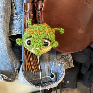 May include: A handmade green crochet creature with orange accents and large black eyes. The whimsical charm is attached to a black keychain. The background includes a brown leather bag and denim jacket.