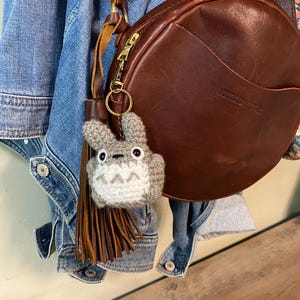 Puede incluir: Un bolso redondo de cuero marrón con cremallera y un borla de cuero marrón unida a un llavero Totoro de ganchillo gris.