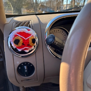 May include: A red, white, and yellow crocheted lion head car air vent decoration. The lion has large, round, yellow eyes and a white mane. The decoration is attached to a car's air vent, with the dashboard and steering wheel visible.