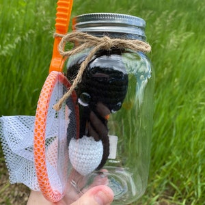 May include: A clear glass jar with a silver lid and a brown twine bow. Inside the jar is a black and white crocheted penguin toy. A bright orange net with a white mesh is next to the jar.