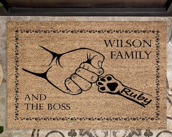 May include: A rectangular coir doormat with a black paw print design and the text "WILSON FAMILY AND THE BOSS Ruby". The mat has a border of paw prints and is placed on a tiled floor.