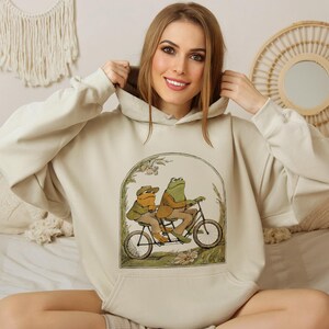 Może przedstawiać: Kremowa bluza z kapturem z grafiką przedstawiającą dwie żaby jadące na rowerze w zielonym i brązowym krajobrazie. Żaby mają na sobie czapki, a rower ma kosz.