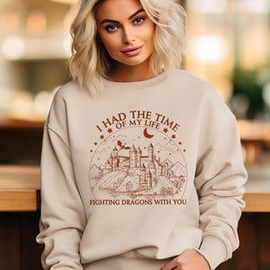 Op de afbeelding: Een beige sweatshirt met een bruine grafische print van een kasteel en een draak. De tekst op de grafiek luidt "Ik had de tijd van mijn leven om draken met je te bevechten".