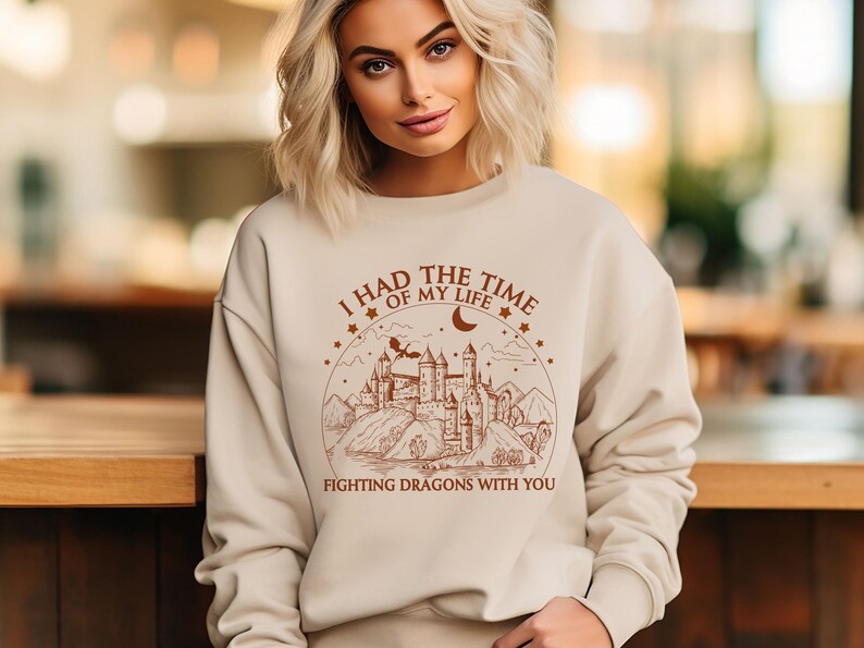 Op de afbeelding: Een beige sweatshirt met een bruine grafische print van een kasteel en een draak. De tekst op de grafiek luidt "Ik had de tijd van mijn leven om draken met je te bevechten".