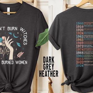 May include: Dark grey heather t-shirt with the text "THEY DIDN'T BURN WITCHES THEY BURNED WOMEN" and a graphic of a hand, flowers, and moons. The back of the shirt lists dates and events related to women's rights.