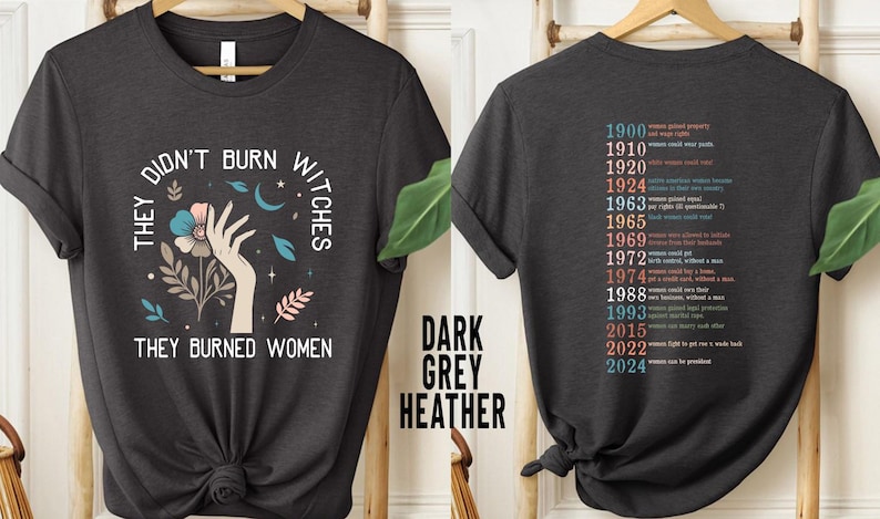 May include: Dark grey heather t-shirt with the text "THEY DIDN'T BURN WITCHES THEY BURNED WOMEN" and a graphic of a hand, flowers, and moons. The back of the shirt lists dates and events related to women's rights.