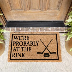以下が含まれることがあります： 「WE'RE PROBABLY AT THE RINK」の黒い文字と、交差したホッケーのスティックとパックが描かれた長方形のドアマット。 マットは天然繊維素材でできており、黒いフレームで囲まれています。