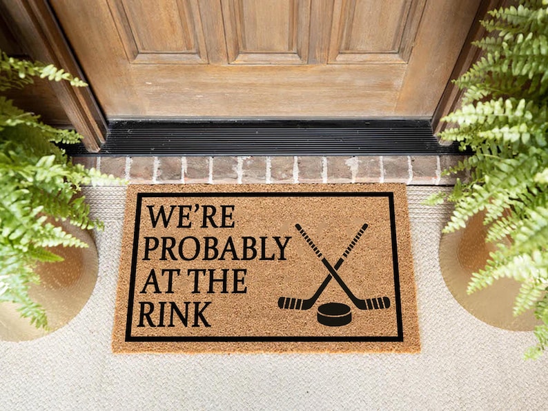 以下が含まれることがあります： 「WE'RE PROBABLY AT THE RINK」の黒い文字と、交差したホッケーのスティックとパックが描かれた長方形のドアマット。 マットは天然繊維素材でできており、黒いフレームで囲まれています。