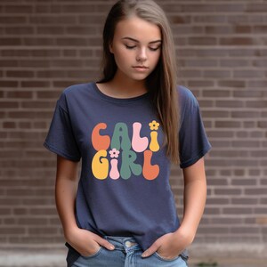 May include: A navy blue t-shirt with a retro-style graphic that says "CALI GIRL" in colorful, wavy letters with flowers.