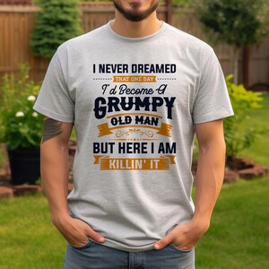 May include: A gray t-shirt with a humorous saying in black and orange text. The text reads "I never dreamed that one day I'd become a grumpy old man but here I am killin' it".