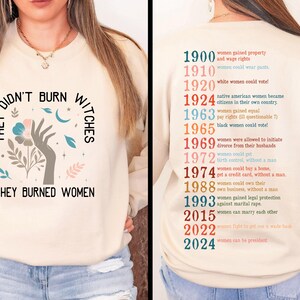 May include: A cream-coloured sweatshirt featuring the text "THEY DIDN'T BURN WITCHES THEY BURNED WOMEN" and a hand holding a flower graphic. The back lists women's rights milestones from 1900 to 2024.