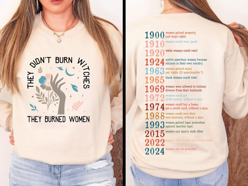 May include: A cream-coloured sweatshirt featuring the text "THEY DIDN'T BURN WITCHES THEY BURNED WOMEN" and a hand holding a flower graphic. The back lists women's rights milestones from 1900 to 2024.