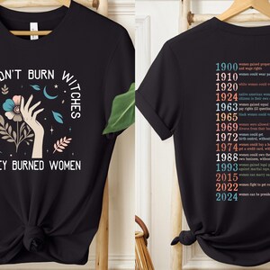 May include: Black t-shirt featuring the text "THEY DIDN'T BURN WITCHES THEY BURNED WOMEN" and a graphic of a hand, flowers, and celestial elements. The back lists women's rights milestones from 1900 to 2024.