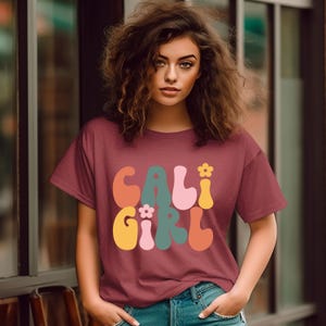 May include: A maroon t-shirt with a retro-style graphic that says "CALI GIRL" in colorful, bubbly letters. The graphic includes small flower accents.