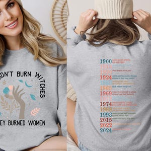 May include: Grey sweatshirt featuring the slogan "THEY DIDN'T BURN WITCHES THEY BURNED WOMEN" with a floral hand graphic. The back displays a timeline of women's rights achievements in various colours.