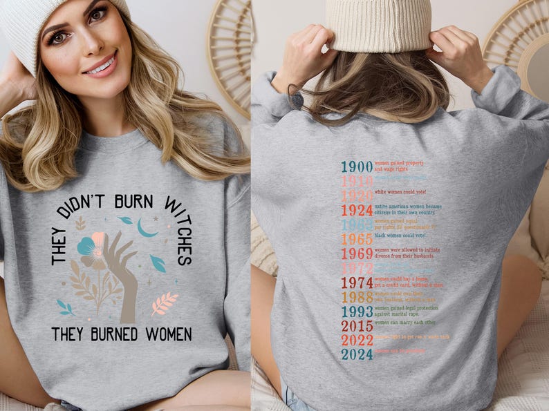 May include: Grey sweatshirt featuring the slogan "THEY DIDN'T BURN WITCHES THEY BURNED WOMEN" with a floral hand graphic. The back displays a timeline of women's rights achievements in various colours.