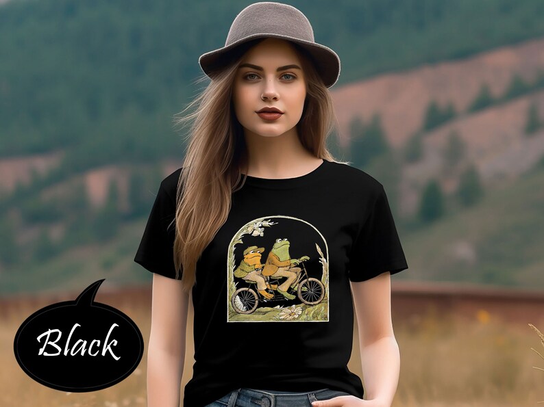 Może przedstawiać: Czarny T-shirt z grafiką przedstawiającą dwie żaby jadące na rowerze w zielonym i brązowym krajobrazie.