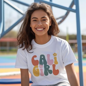 May include: A white t-shirt with a colorful graphic that says "CALI GIRL" in a retro font. The graphic features flowers and different shades of orange, pink, yellow, green, and brown.