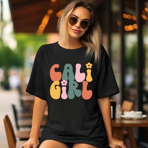 May include: Black oversized t-shirt with a retro-style graphic that says "CALI GIRL" in colorful, wavy letters with small flower accents.