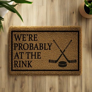 以下が含まれることがあります： 茶色の背景と黒い縁取りの長方形のドアマット。「WE'RE PROBABLY AT THE RINK」の文字が黒で印刷され、ホッケーのスティックとパックが交差しています。白いスニーカーがマットの隣にあります。