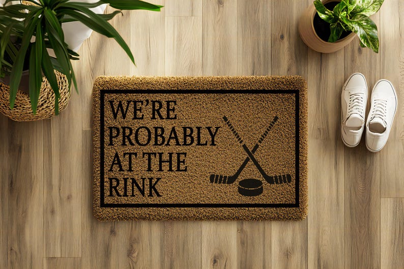 以下が含まれることがあります： 茶色の背景と黒い縁取りの長方形のドアマット。「WE'RE PROBABLY AT THE RINK」の文字が黒で印刷され、ホッケーのスティックとパックが交差しています。白いスニーカーがマットの隣にあります。