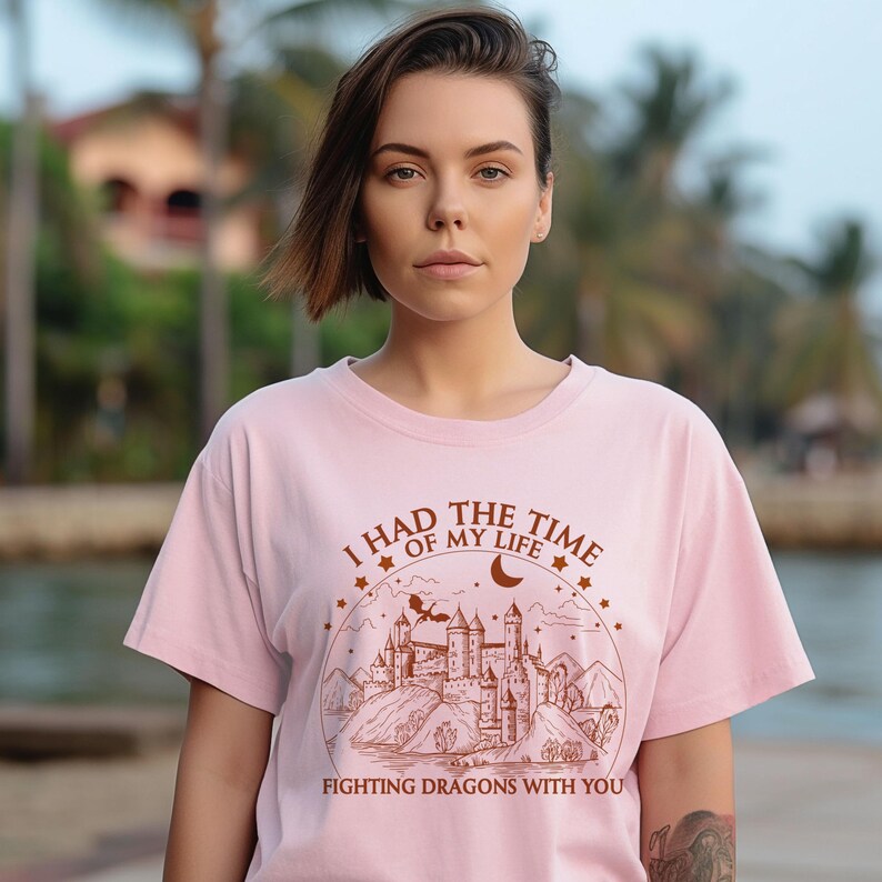 Op de afbeelding: Roze T-shirt met een bruine afbeelding van een kasteel, maan en sterren. De tekst op het shirt luidt "Ik had de tijd van mijn leven om draken met je te bevechten."