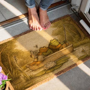 May include: A rectangular doormat featuring a whimsical illustration of two frogs in a boat on a pond. One frog is reading a book, while the other is fishing. The mat has a brown border and a light beige background.