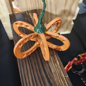 Puede incluir: Una escultura decorativa de calabaza construida con herraduras de metal pintadas de naranja dispuestas en forma esférica. La calabaza presenta un tallo de metal retorcido pintado de verde unido a la parte superior central de la estructura.