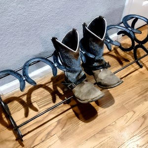 Könnte beinhalten: Ein schwarzer Metall-Stiefelständer mit Hufeisen-Design hält zwei Paar Cowboy-Stiefel. Die Stiefel sind braun und grau mit dekorativen Nähten und Riemen. Der Ständer steht auf einem Holzboden.