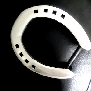 May include: A white horseshoe featuring a U-shaped design with six evenly spaced square nail holes along its curved surface. The object is presented against a dark, solid background, highlighting its smooth, matte finish and distinct arched form.