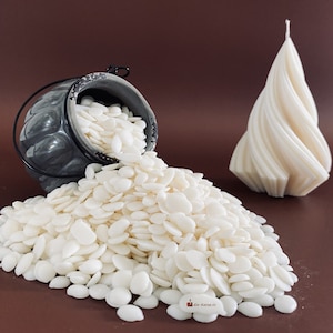 May include: A gray ceramic pot with a black metal handle is overflowing with white wax flakes. A white, spiral-shaped candle sits on a brown surface.