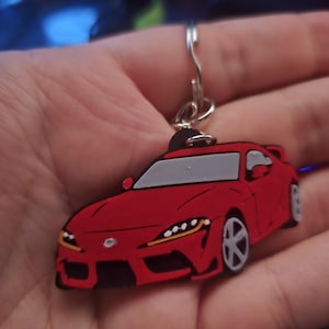 May include: A red car-shaped keyring with a silver ring. The keyring features a stylized design of a sports car with grey windows and black accents. The car has yellow and white headlights.