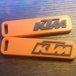 May include: Two orange keychains featuring the black KTM logo. The keychains are rectangular with rounded edges and a hole for a keyring. The KTM logo is a stylized wordmark.