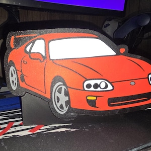 Op de afbeelding: Een rode sportwagen uitgesneden met zwarte afwerking en witte ramen. De auto heeft zilveren wielen en een achterspoiler. Het is een gestileerde illustratie van een Toyota Supra.